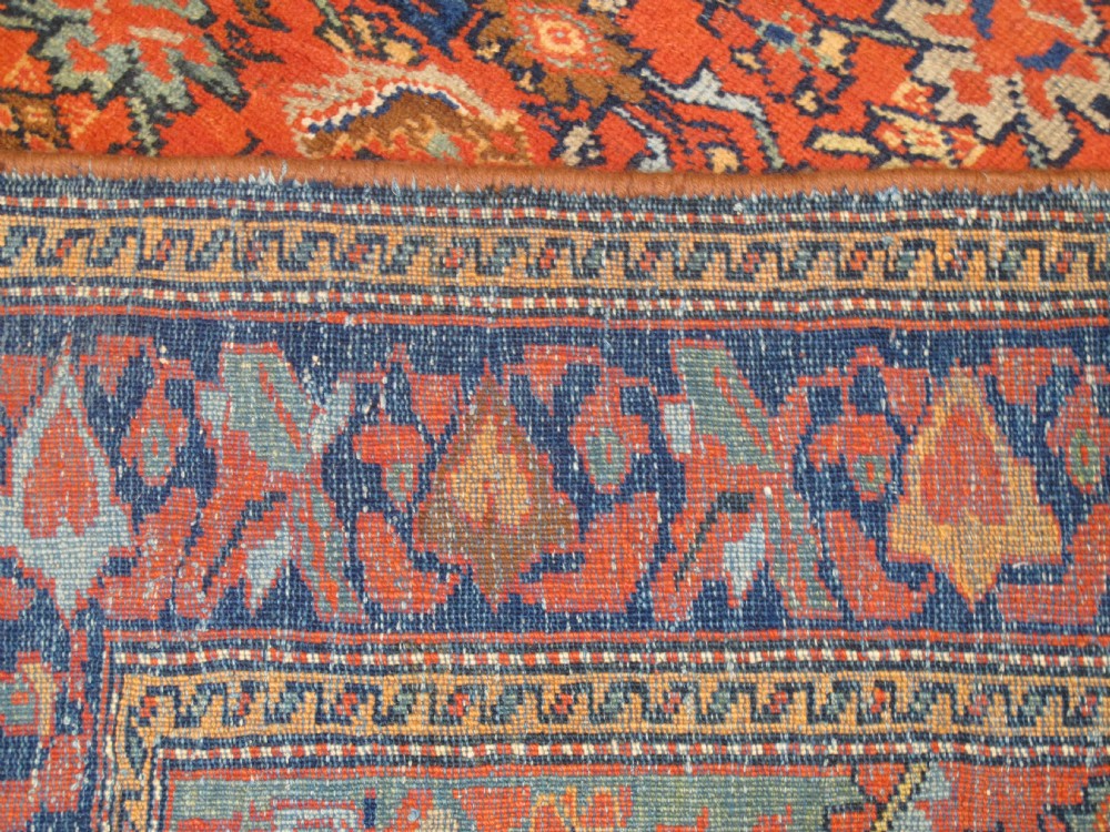 An antique hand-knotted West Persian wool rug featuring a central rust red field with geometric motifs and a dark blue patterned border. View VI