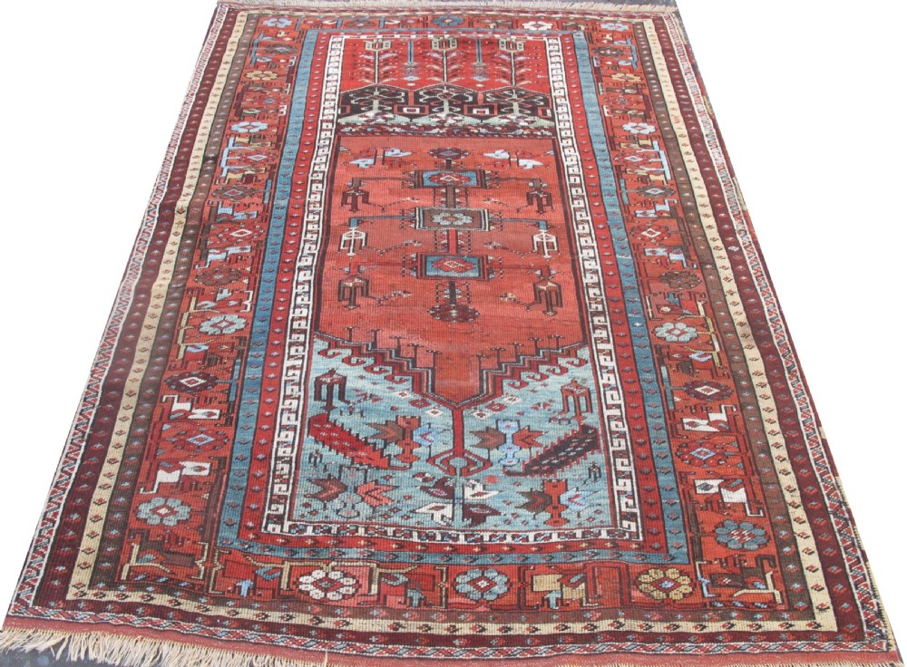 An antique Anatolian village rug featuring a red central field with geometric motifs in light blue, cream, and brown, framed by patterned multi-tiered borders.