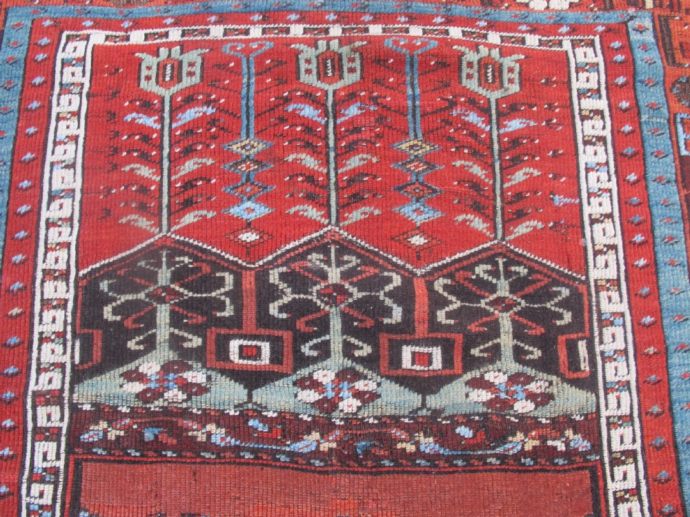 An antique Anatolian village rug featuring a red central field with geometric motifs in light blue, cream, and brown, framed by patterned multi-tiered borders. View II