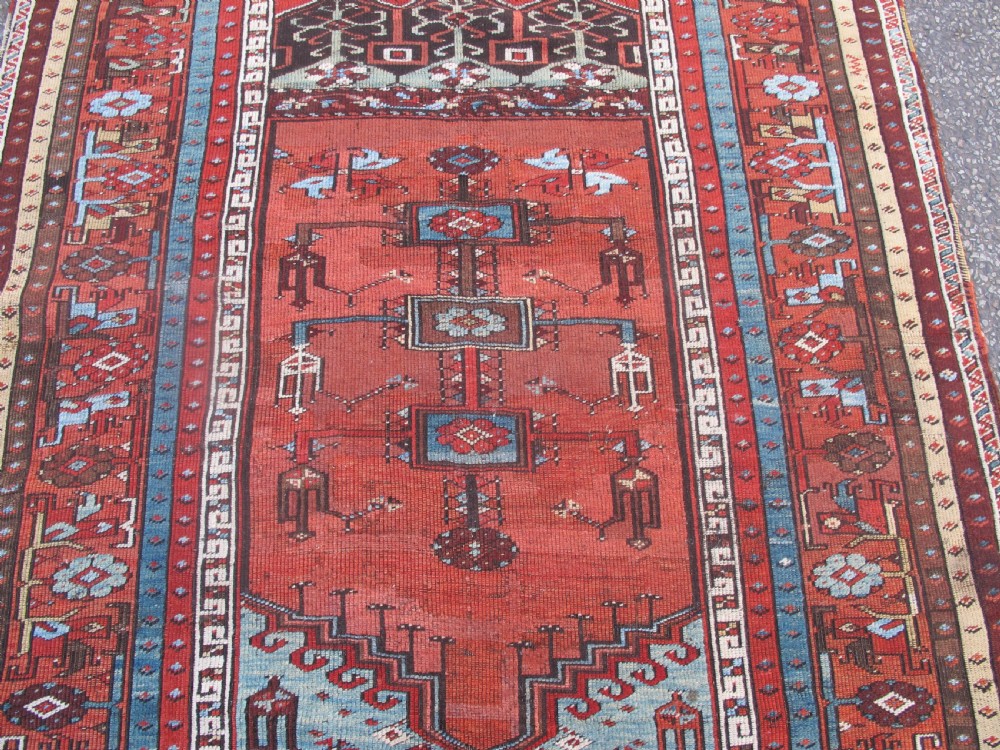 An antique Anatolian village rug featuring a red central field with geometric motifs in light blue, cream, and brown, framed by patterned multi-tiered borders. View III