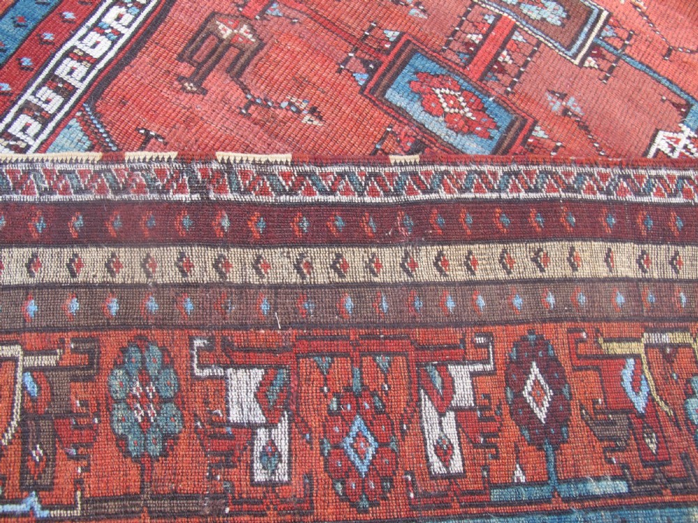An antique Anatolian village rug featuring a red central field with geometric motifs in light blue, cream, and brown, framed by patterned multi-tiered borders. View IV