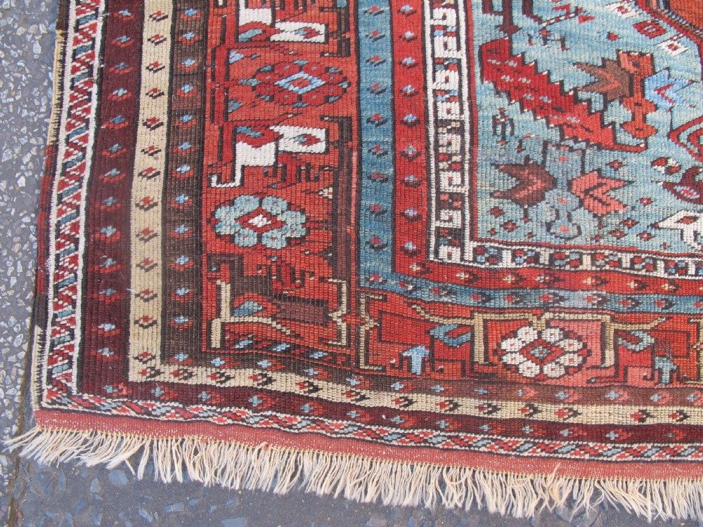 An antique Anatolian village rug featuring a red central field with geometric motifs in light blue, cream, and brown, framed by patterned multi-tiered borders. View V