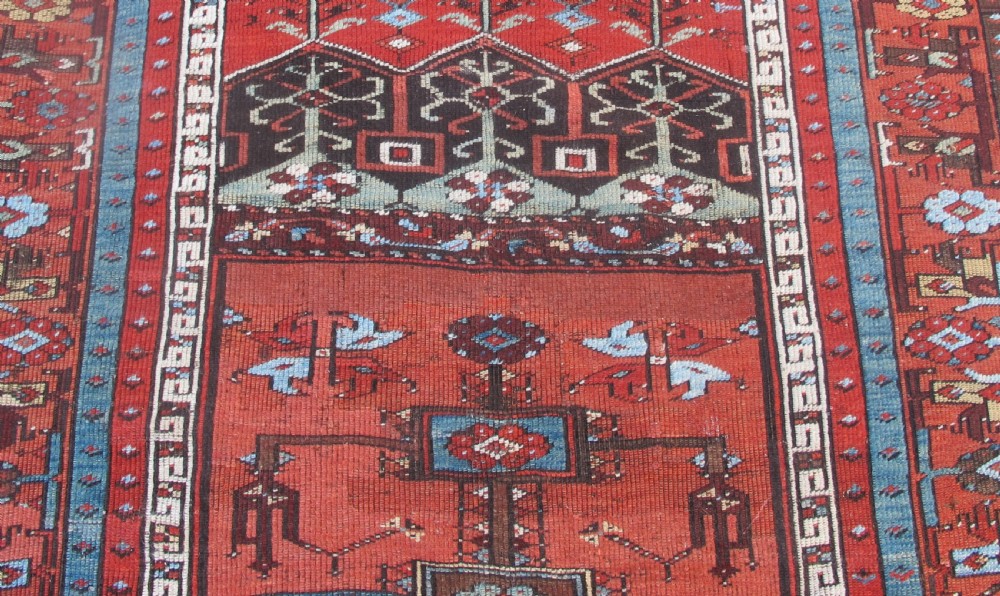 An antique Anatolian village rug featuring a red central field with geometric motifs in light blue, cream, and brown, framed by patterned multi-tiered borders. View VI