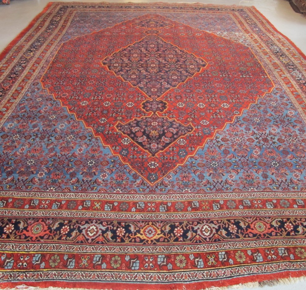 A large antique Persian Bidjar carpet featuring a central diamond medallion, red field, blue spandrels, and intricate floral borders.