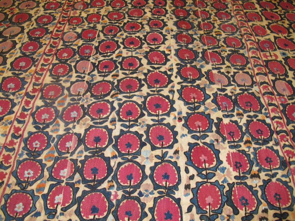 A large rectangular antique suzani textile featuring repeating circular red floral motifs framed by a decorative floral border on an off-white ground. View V