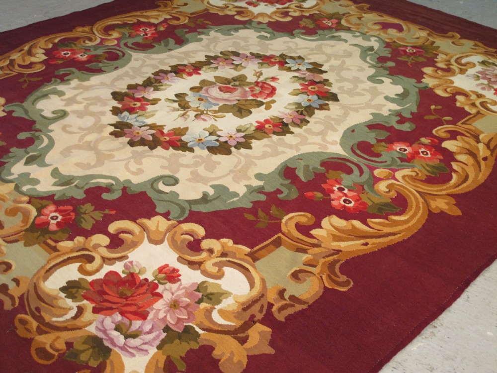 A rectangular flatweave Aubusson wool carpet featuring a central floral medallion surrounded by acanthus scrollwork cartouches on a deep red ground. View IV