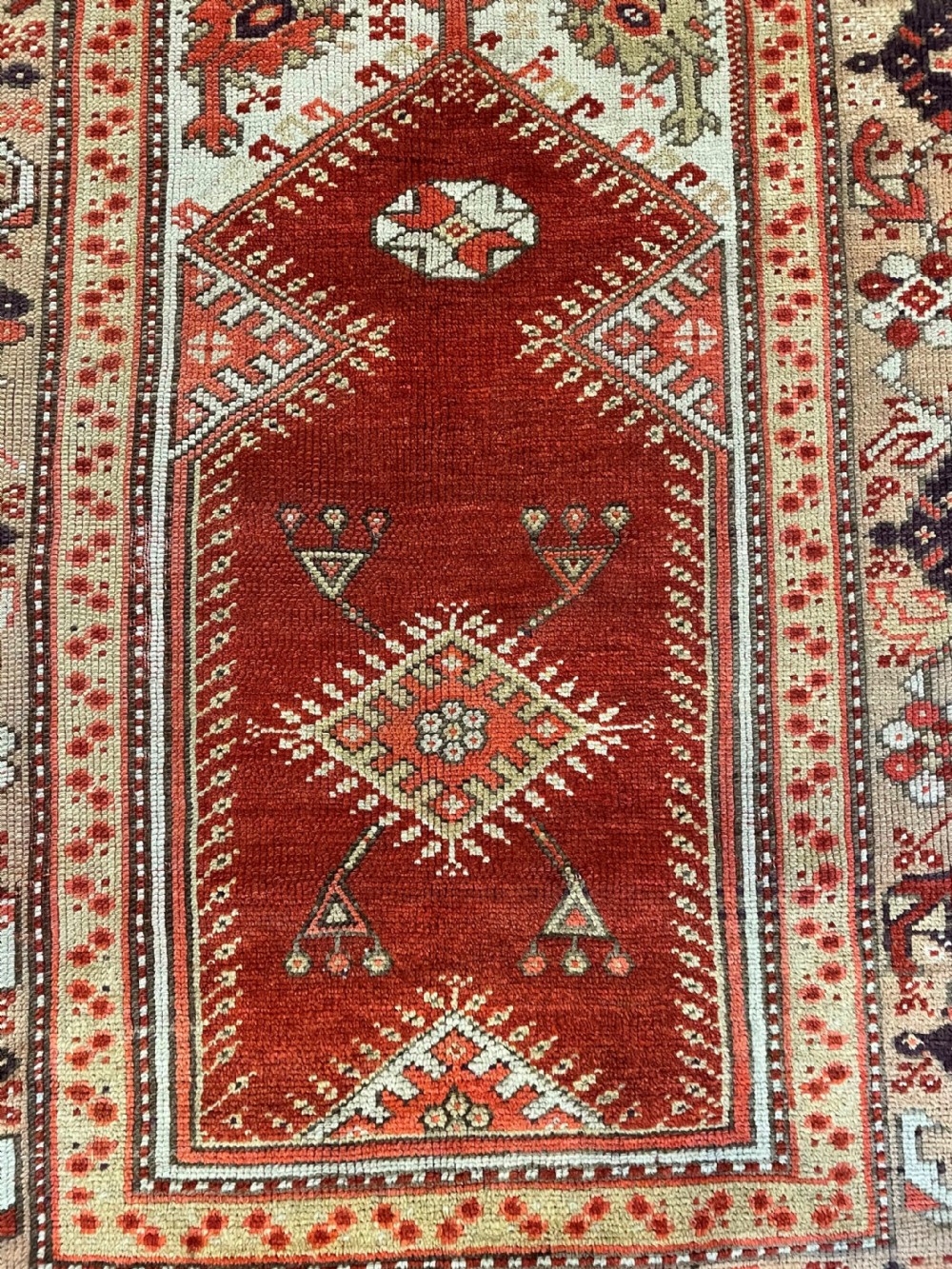 A rectangular hand-knotted Anatolian Melas wool rug featuring a red central pointed mihrab field framed by broad geometric patterned borders in cream, ochre, and dark brown. View VII