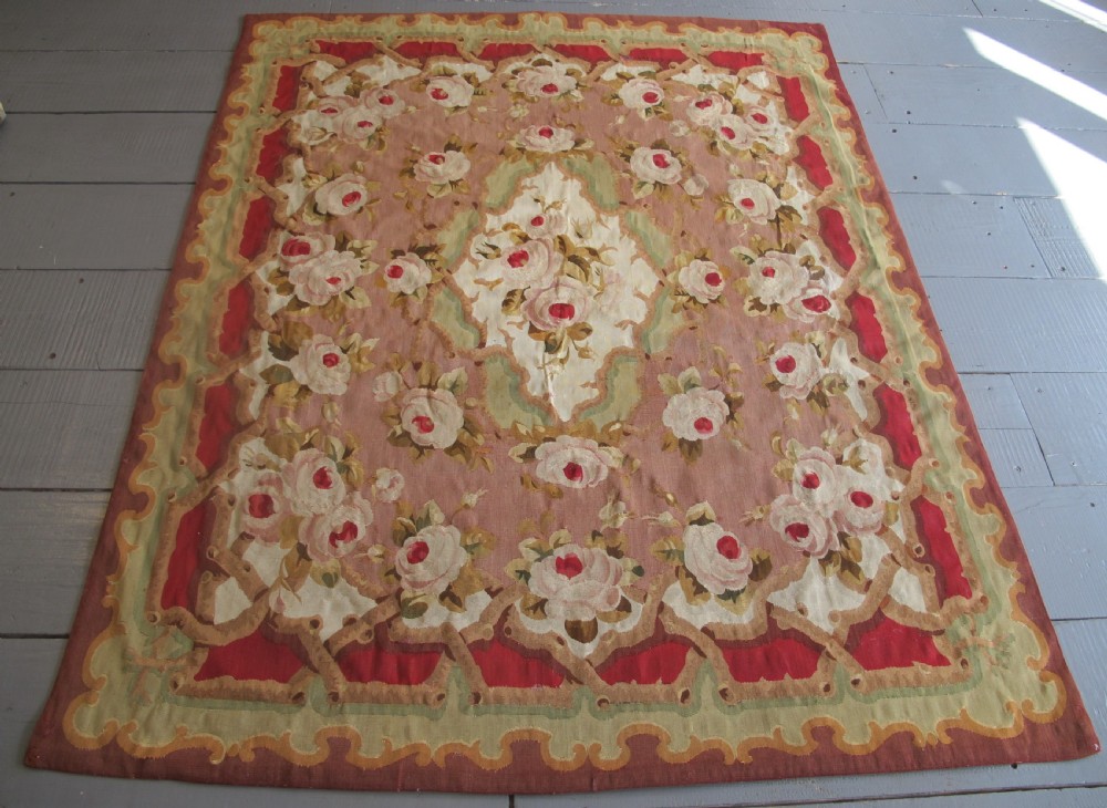 A rectangular antique French Aubusson flatweave wool rug featuring a central floral medallion surrounded by rose motifs, scrollwork borders, and shades of pink, red, green, and cream.