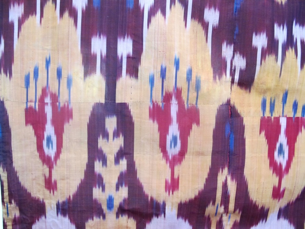 A rectangular handwoven silk ikat textile panel displaying repeating floral and botanical motifs in shades of burgundy, magenta, pink, and ochre yellow. View II