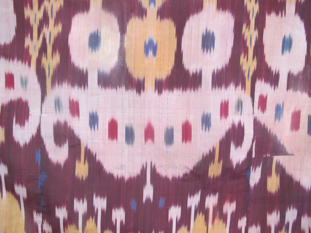 A rectangular handwoven silk ikat textile panel displaying repeating floral and botanical motifs in shades of burgundy, magenta, pink, and ochre yellow. View III