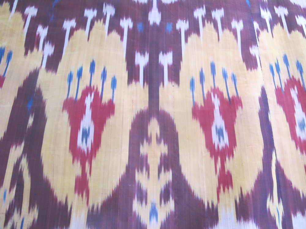 A rectangular handwoven silk ikat textile panel displaying repeating floral and botanical motifs in shades of burgundy, magenta, pink, and ochre yellow. View V
