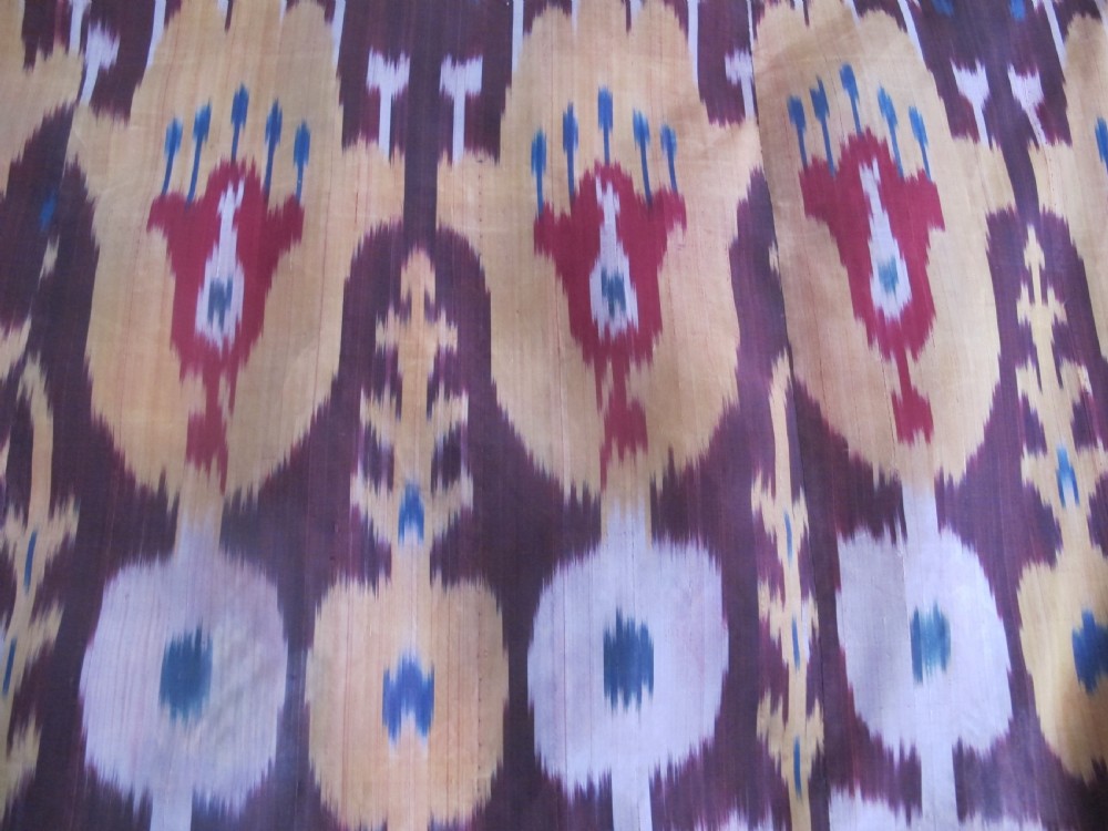 A rectangular handwoven silk ikat textile panel displaying repeating floral and botanical motifs in shades of burgundy, magenta, pink, and ochre yellow. View VI