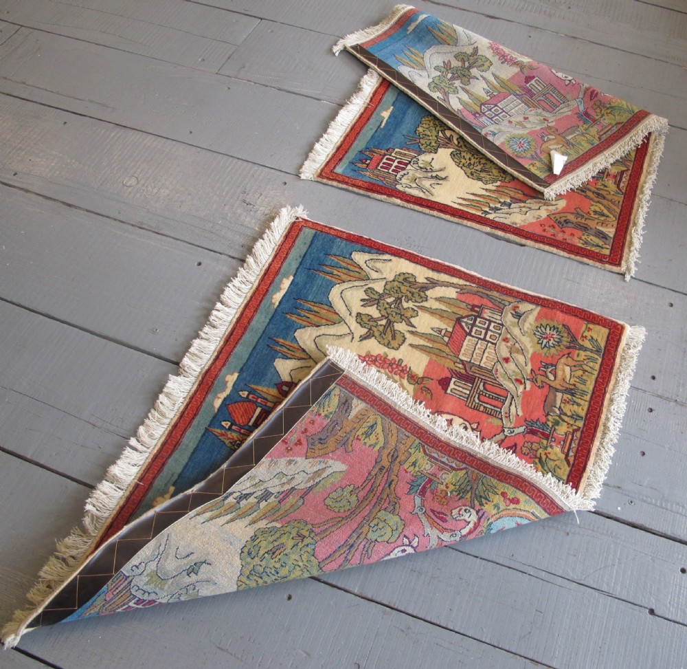 A pair of small Persian Kashan pictorial wool rugs featuring woven architectural scenes, trees, and animals set against terracotta red, cream, and navy blue backgrounds. View X