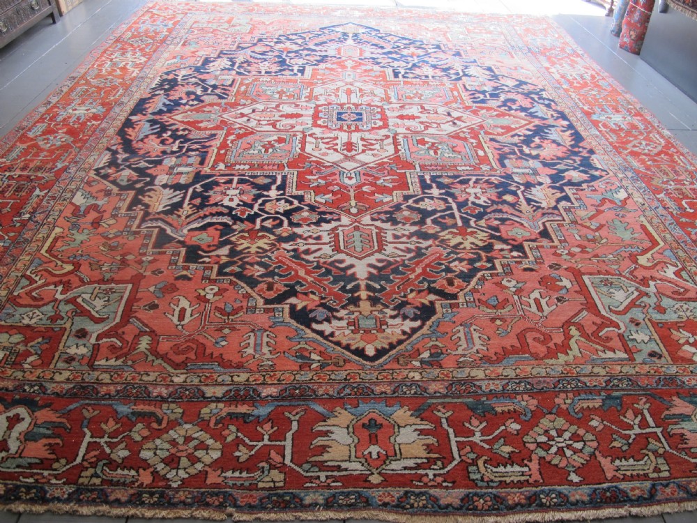 A large antique Persian Heriz carpet featuring a central geometric medallion surrounded by stylized floral motifs in terracotta, navy blue, and cream tones.