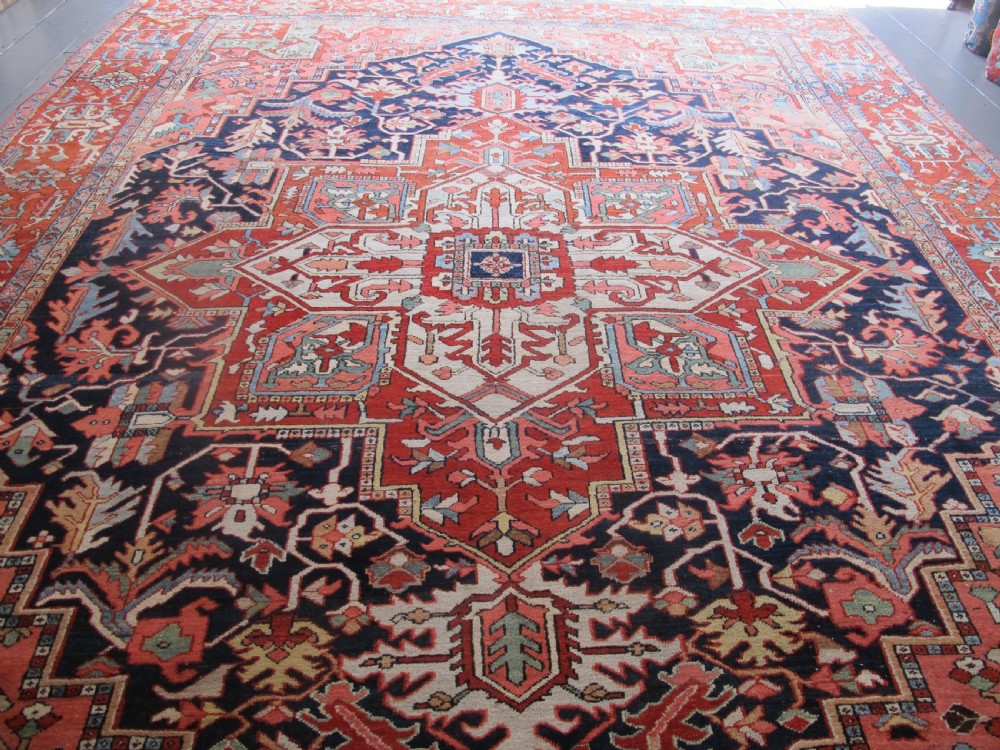 A large antique Persian Heriz carpet featuring a central geometric medallion surrounded by stylized floral motifs in terracotta, navy blue, and cream tones. View II