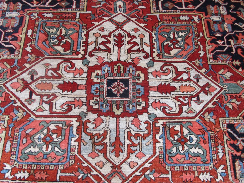 A large antique Persian Heriz carpet featuring a central geometric medallion surrounded by stylized floral motifs in terracotta, navy blue, and cream tones. View V