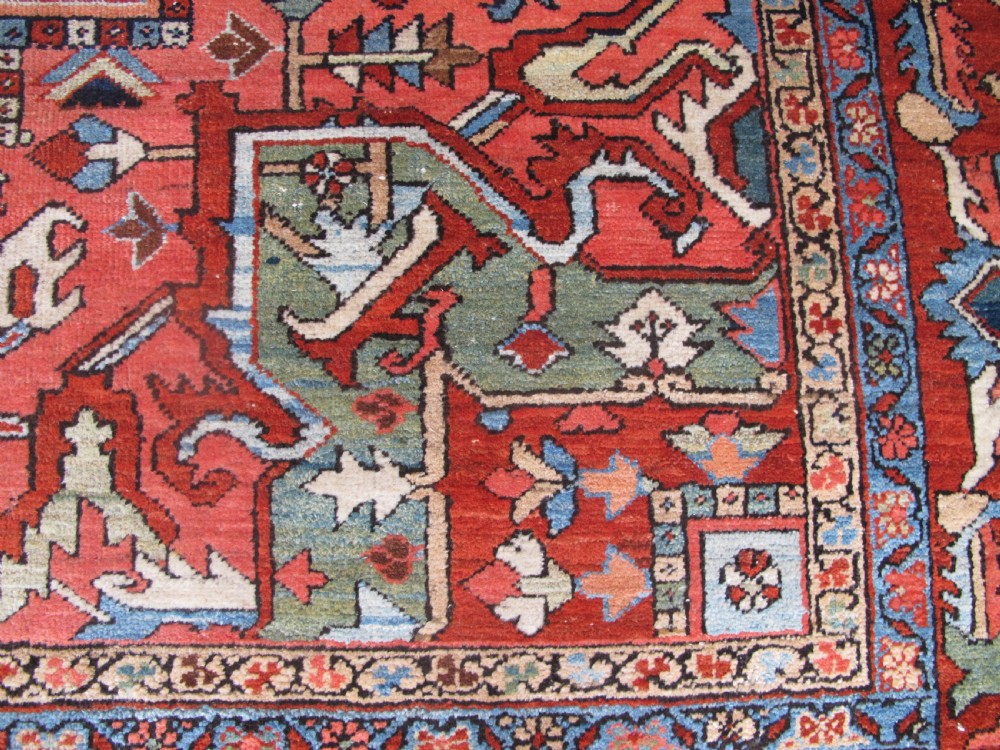 A large antique Persian Heriz carpet featuring a central geometric medallion surrounded by stylized floral motifs in terracotta, navy blue, and cream tones. View VI