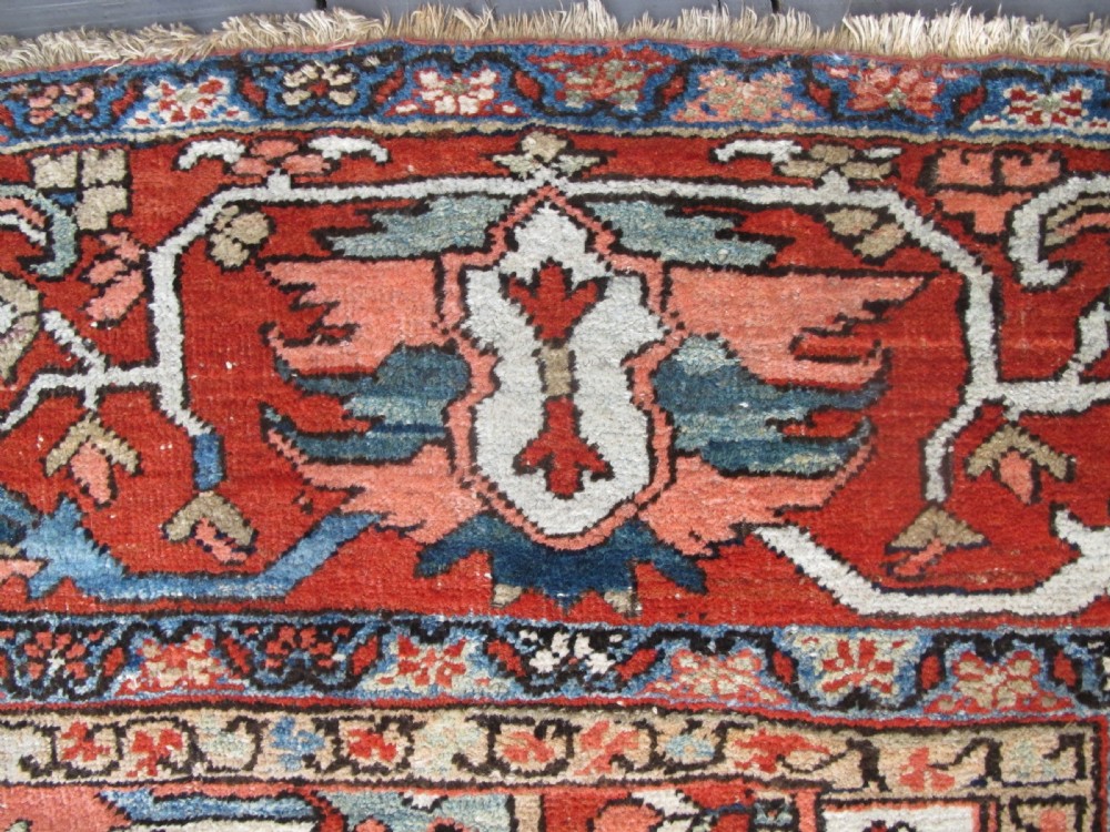 A large antique Persian Heriz carpet featuring a central geometric medallion surrounded by stylized floral motifs in terracotta, navy blue, and cream tones. View VII