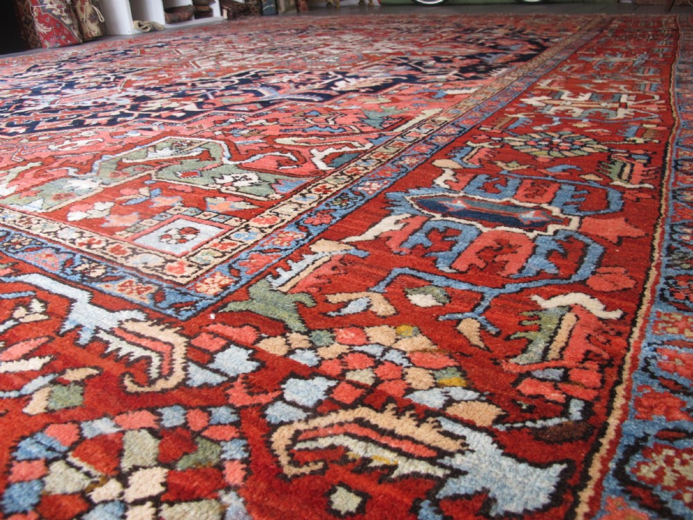 A large antique Persian Heriz carpet featuring a central geometric medallion surrounded by stylized floral motifs in terracotta, navy blue, and cream tones. View X