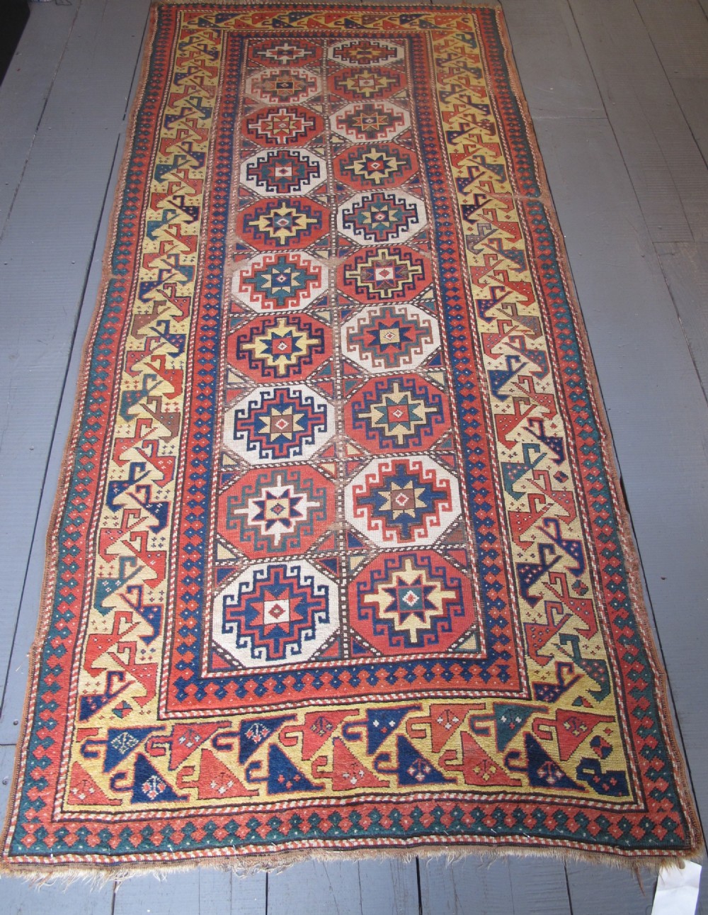 An antique hand-knotted wool Caucasian rug runner featuring a central column of stepped octagonal Memling guls on a red background bordered by geometric patterns.