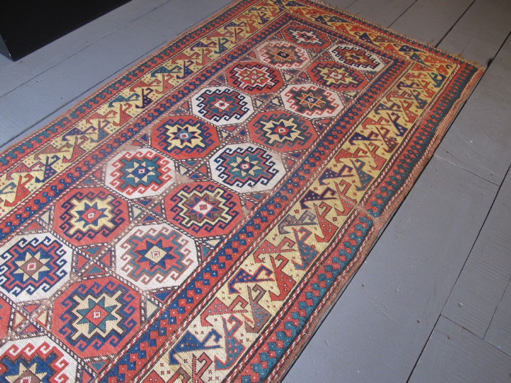 An antique hand-knotted wool Caucasian rug runner featuring a central column of stepped octagonal Memling guls on a red background bordered by geometric patterns. View II