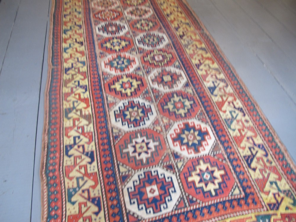 An antique hand-knotted wool Caucasian rug runner featuring a central column of stepped octagonal Memling guls on a red background bordered by geometric patterns. View III