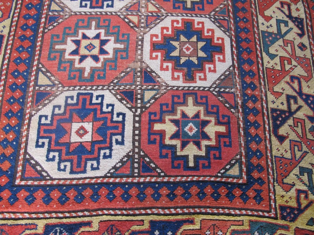 An antique hand-knotted wool Caucasian rug runner featuring a central column of stepped octagonal Memling guls on a red background bordered by geometric patterns. View IV