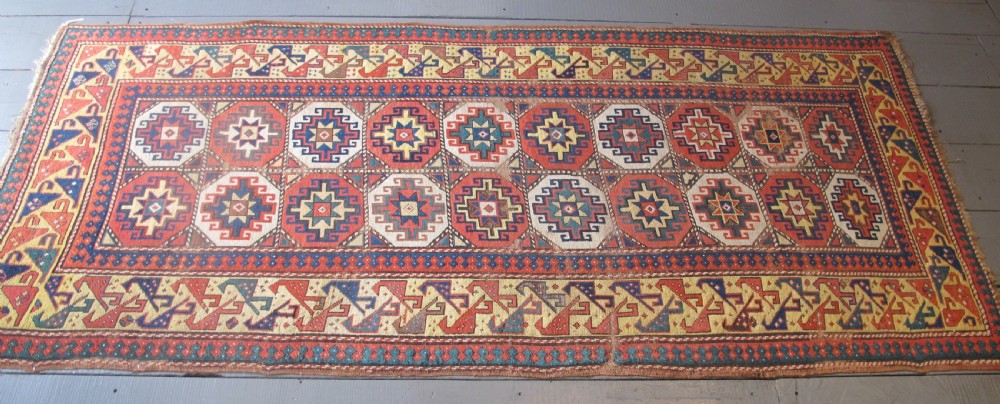 An antique hand-knotted wool Caucasian rug runner featuring a central column of stepped octagonal Memling guls on a red background bordered by geometric patterns. View X