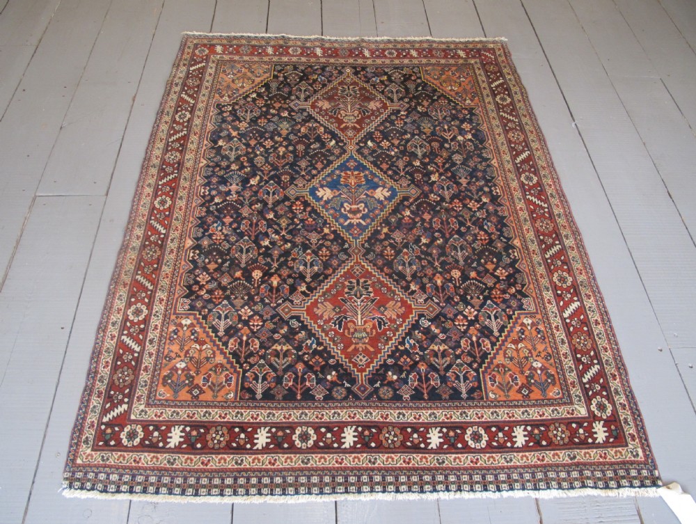 A hand-knotted Persian Qashqai wool rug featuring a dark navy field with central diamond medallions, geometric floral motifs, and a multi-tiered decorative border.