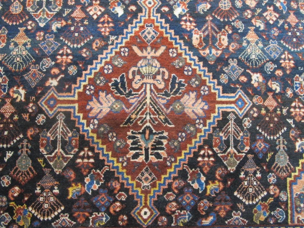 A hand-knotted Persian Qashqai wool rug featuring a dark navy field with central diamond medallions, geometric floral motifs, and a multi-tiered decorative border. View IV