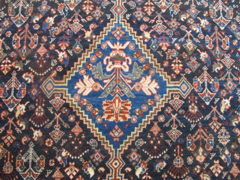 A hand-knotted Persian Qashqai wool rug featuring a dark navy field with central diamond medallions, geometric floral motifs, and a multi-tiered decorative border. View V