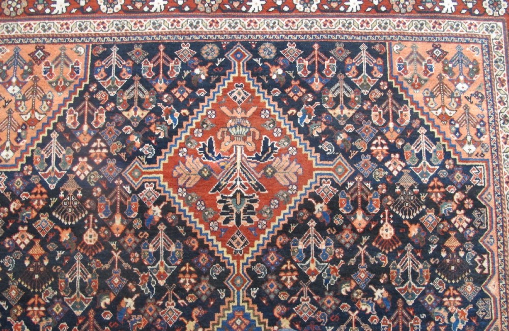 A hand-knotted Persian Qashqai wool rug featuring a dark navy field with central diamond medallions, geometric floral motifs, and a multi-tiered decorative border. View VI