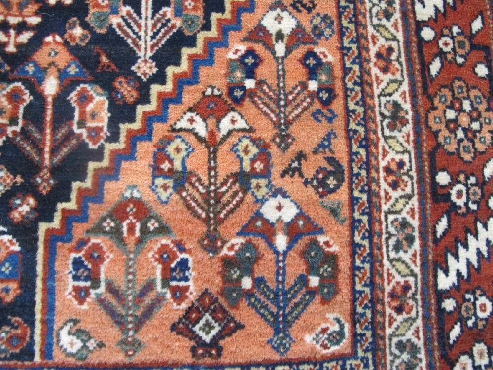 A hand-knotted Persian Qashqai wool rug featuring a dark navy field with central diamond medallions, geometric floral motifs, and a multi-tiered decorative border. View VII