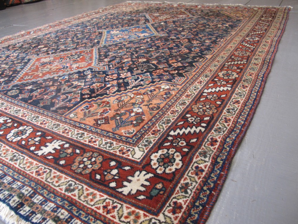 A hand-knotted Persian Qashqai wool rug featuring a dark navy field with central diamond medallions, geometric floral motifs, and a multi-tiered decorative border. View X