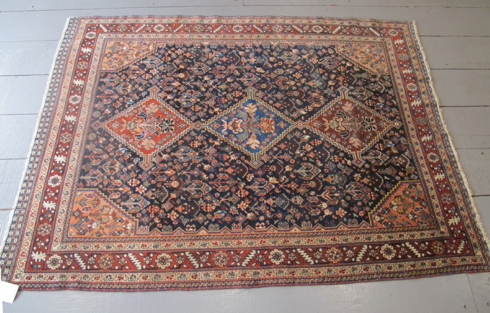 A hand-knotted Persian Qashqai wool rug featuring a dark navy field with central diamond medallions, geometric floral motifs, and a multi-tiered decorative border. View XII