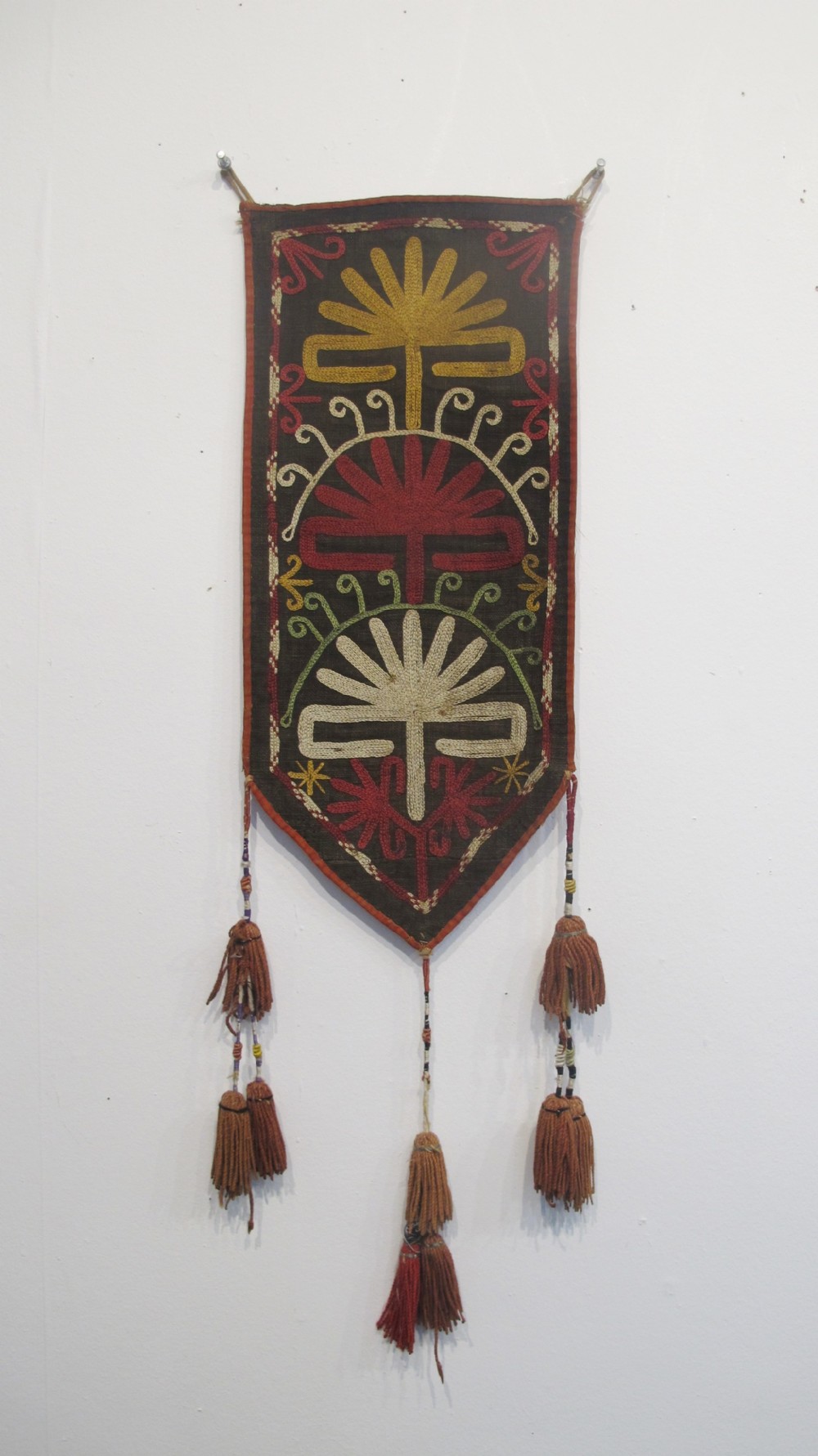 An antique Uzbek vertical silk embroidered textile amulet featuring yellow, red, and cream palmette motifs on a dark brown ground with hanging tassels.