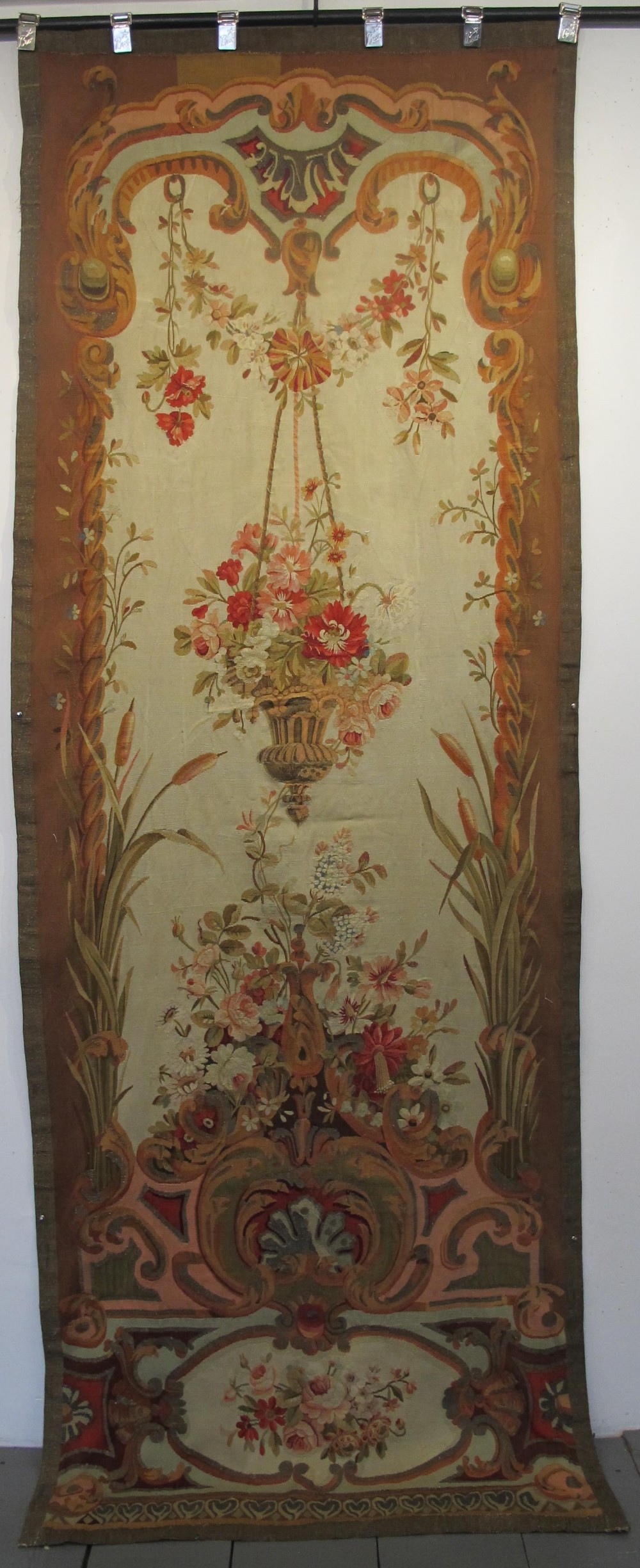 A tall, narrow 19th-century French Aubusson woven tapestry featuring a hanging floral basket, urn motifs, and rocaille border designs on a beige ground.