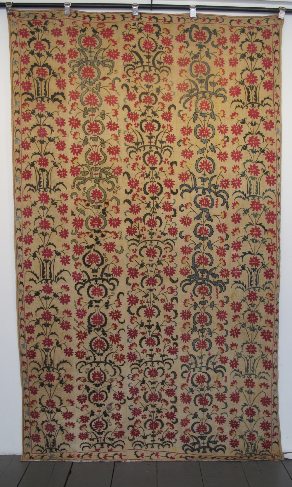 A large rectangular Central Asian suzani textile featuring repeating vertical columns of red embroidered floral motifs on a natural beige cotton substrate.