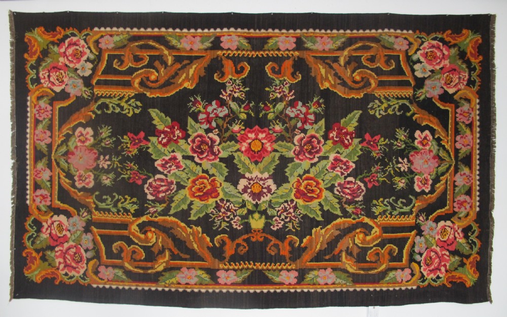 A large rectangular handwoven flatweave rug featuring vibrant floral bouquets and scrollwork borders on a dark black field.