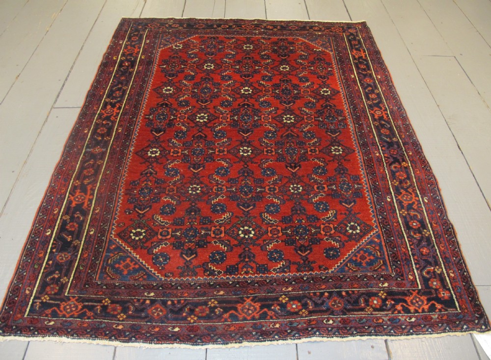 An antique West Persian Melayir wool rug featuring a deep red central field filled with repeating geometric floral motifs within a dark navy multi-tiered border.