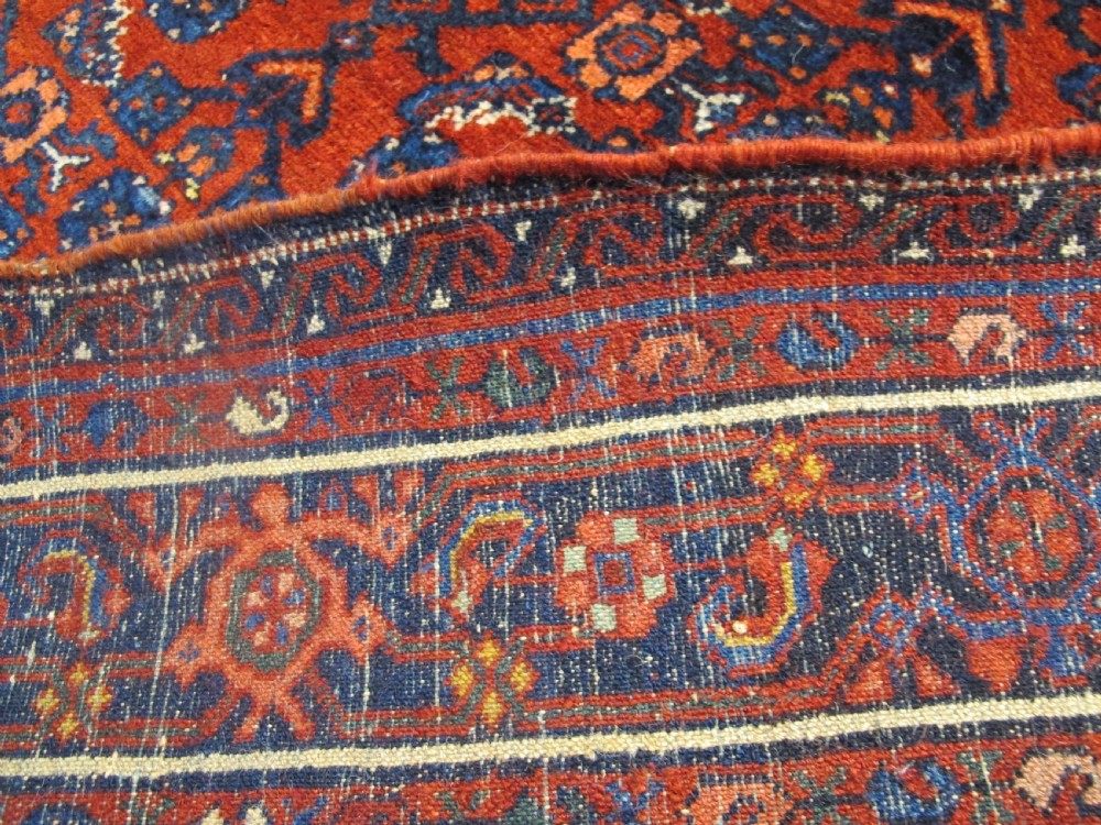An antique West Persian Melayir wool rug featuring a deep red central field filled with repeating geometric floral motifs within a dark navy multi-tiered border. View IV