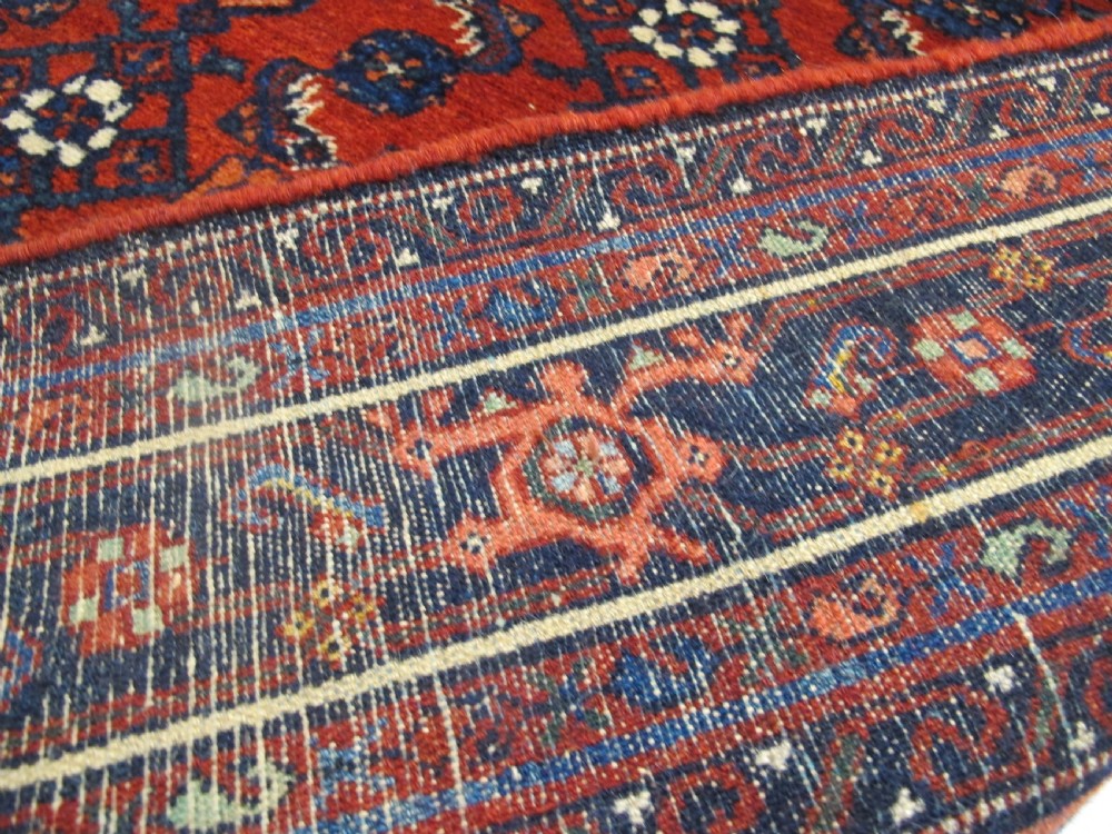 An antique West Persian Melayir wool rug featuring a deep red central field filled with repeating geometric floral motifs within a dark navy multi-tiered border. View V