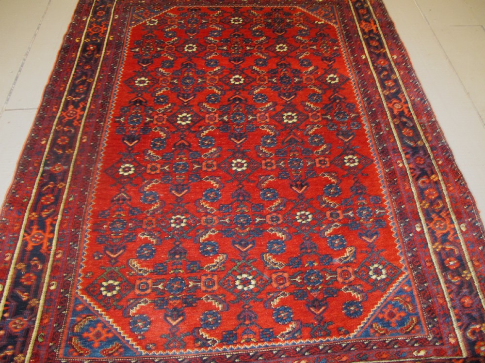 An antique West Persian Melayir wool rug featuring a deep red central field filled with repeating geometric floral motifs within a dark navy multi-tiered border. View VI