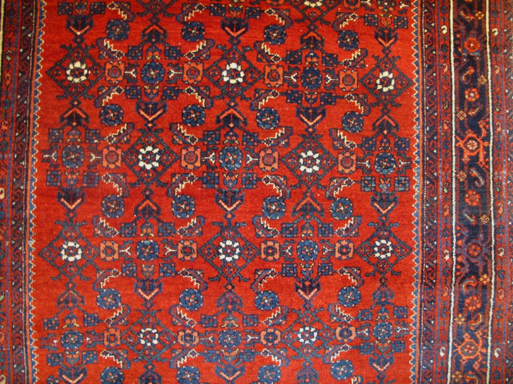 An antique West Persian Melayir wool rug featuring a deep red central field filled with repeating geometric floral motifs within a dark navy multi-tiered border. View IX