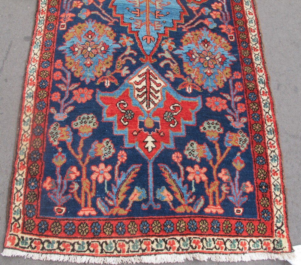A long, narrow hand-knotted Persian wool runner featuring intricate geometric floral medallions in shades of navy blue, terracotta red, and sky blue along a dark central field. View II