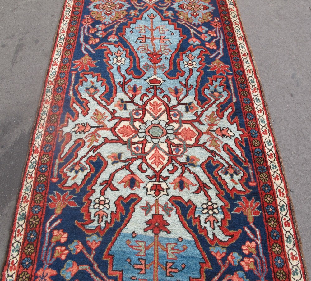 A long, narrow hand-knotted Persian wool runner featuring intricate geometric floral medallions in shades of navy blue, terracotta red, and sky blue along a dark central field. View IV