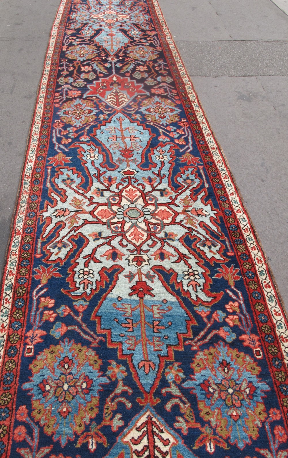 A long, narrow hand-knotted Persian wool runner featuring intricate geometric floral medallions in shades of navy blue, terracotta red, and sky blue along a dark central field. View VII