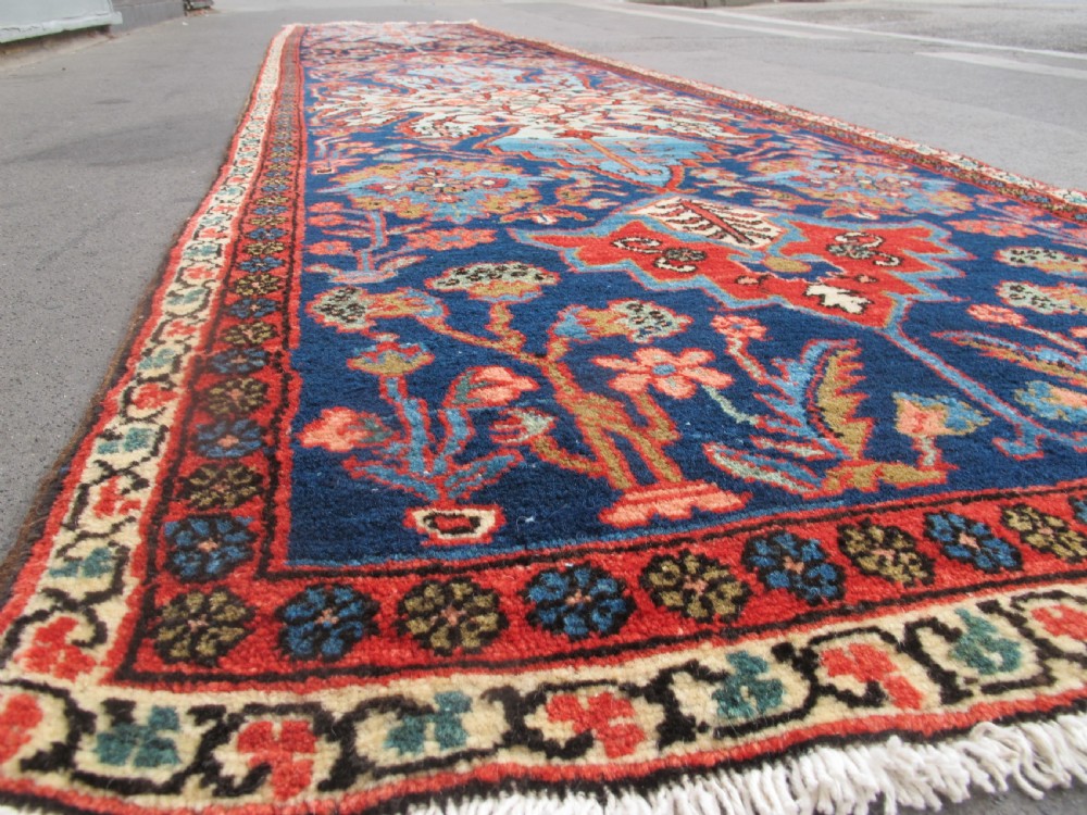 A long, narrow hand-knotted Persian wool runner featuring intricate geometric floral medallions in shades of navy blue, terracotta red, and sky blue along a dark central field. View VIII