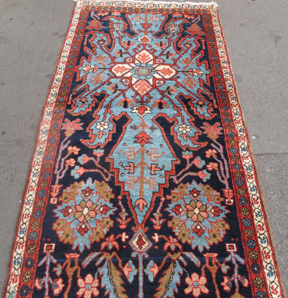 A long, narrow hand-knotted Persian wool runner featuring intricate geometric floral medallions in shades of navy blue, terracotta red, and sky blue along a dark central field. View IX
