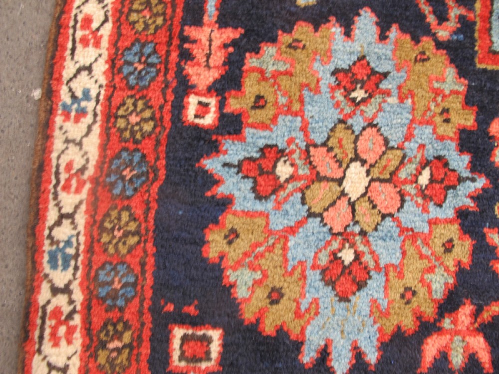 A long, narrow hand-knotted Persian wool runner featuring intricate geometric floral medallions in shades of navy blue, terracotta red, and sky blue along a dark central field. View XI